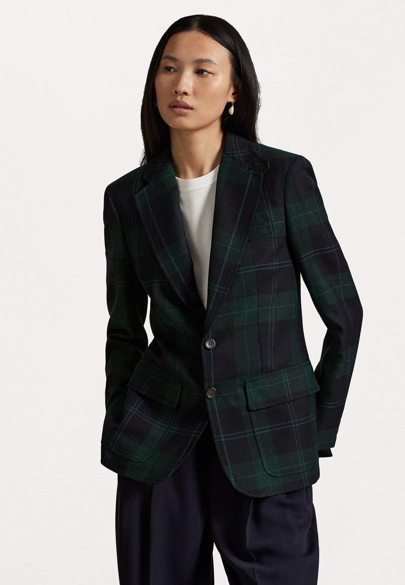 Žena s dlhými tmavými vlasmi má na sebe zeleno-námorníkovo pruhovaný blejzer cez biely top a voľné čierne nohavice, stojí na bielom pozadí.