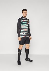 Langarm sporttrui met zwarte en groene horizontale strepen, ritsnek, zwarte shorts, zwarte sokken en zwarte voetbalschoenen met zilveren accenten.
