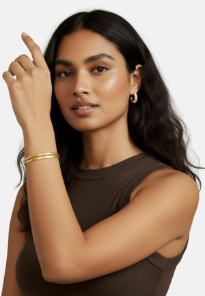 Bracciale in metallo dorato con tre fasce parallele; indossato su un braccio. Il modello ha capelli lunghi e scuri e indossa un top aderente scuro.