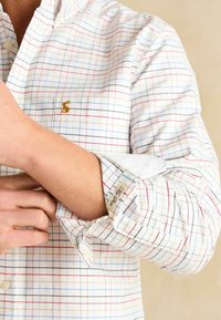 Biela košeľa na gombíky s mriežkovaným vzorom v červenej, modrej a čiernej farbe. Má zviazané rukávy a vyšívané logo na hrudi.