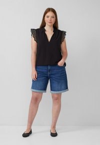 Blouse noire avec des manches bordées de dentelle, design en V ; associée à un short en denim bleu, avec des poignets retroussés. Des chaussures plates noires complètent la tenue.