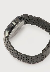 Μαύρη μπρασελέ από μεταλλικούς κρίκους με πλευρικά κουμπιά και χαρακτηριστικό κούμπωμα, εμφανιζόμενη σε κυκλική μορφή από πλάγια γωνία.