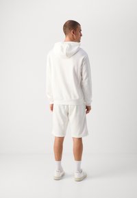 Completo in bianco con felpa e pantaloncini, caratterizzato da cappuccio, polsini elasticizzati e vestibilità comoda. Abbinato a sneakers bianche e calze.