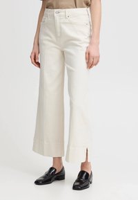 Pantaloni ampi di colore crema con vita alta, tasche frontali e spacchi laterali all'orlo, realizzati in tessuto di cotone. Mocassini neri in vista.