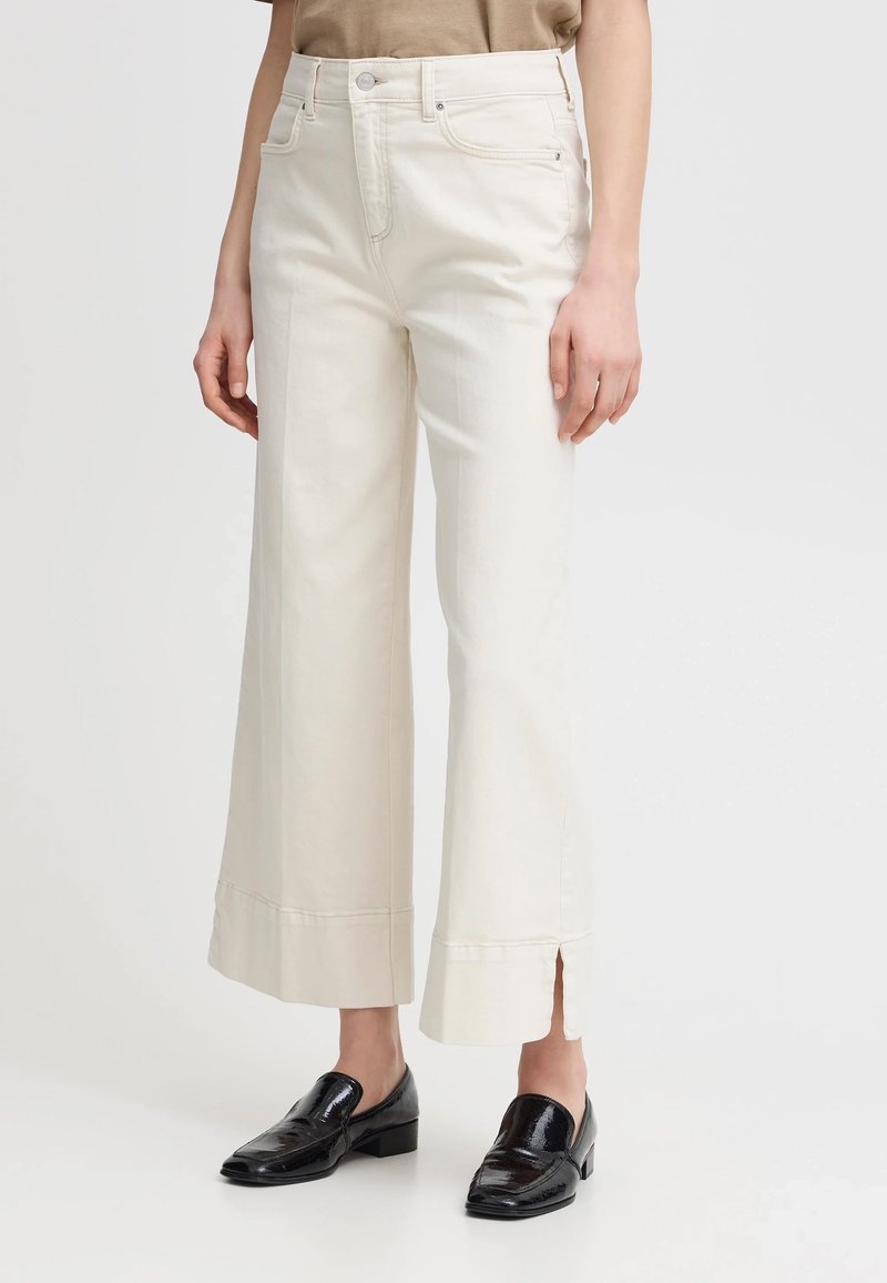 Pantaloni ampi di colore crema con vita alta, tasche frontali e spacchi laterali all'orlo, realizzati in tessuto di cotone. Mocassini neri in vista.