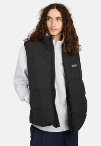 Schwarze isolierte Pufferweste mit Reißverschluss, hohem Kragen und horizontalem Steppmuster, getragen über einem hellgrauen Kapuzenpullover.