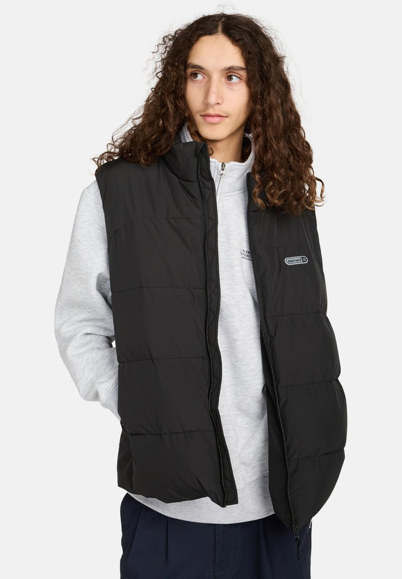 Schwarze isolierte Pufferweste mit Reißverschluss, hohem Kragen und horizontalem Steppmuster, getragen über einem hellgrauen Kapuzenpullover.