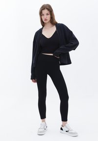 Sudadera negra corta con cremallera, camiseta ajustada negra y leggins negros de talle alto; combinados con zapatillas blancas con detalles negros.