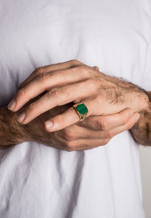 Gold ring featuring a rectangular green stone with vertical lines. The hands are lightly hairy, resting against a white shirt fabric.