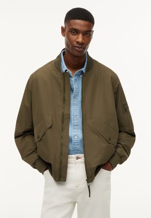 Homme portant une veste bomber vert olive par-dessus une chemise bleu clair boutonnée et un pantalon blanc, les mains dans les poches, sur un fond uni.