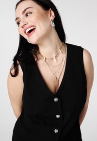 Gilet noir texturé avec un col en V profond et des boutons en forme de cœur argentés ; colliers superposés en or avec des accents blancs ; grandes boucles d'oreilles en argent.