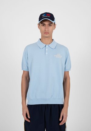 Jonge man die een lichtblauw poloshirt en een donkere pet met roodblauw ontwerp draagt, staande tegen een effen witte achtergrond.