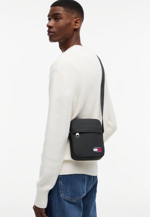 Vīrietis baltā džemperī un zilos džinsos, nēsājot melnu Tommy Jeans plecu somu pāri plecam.