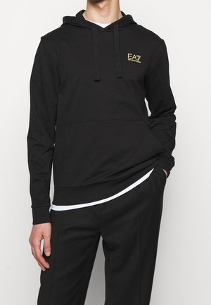 Mężczyzna ubrany w czarną bluzę z kapturem EA7 Emporio Armani z przednią kieszenią i czarne spodnie, ręka w kieszeni, na jednolitym białym tle.