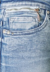 Vaaleansiniset farkut lähikuvassa, jossa on etutasku, pieni vetoketju, niittejä ja näkyvää kankaan rakennetta.