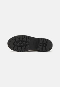 Suela de zapato de goma negra con un patrón de relieve profundo, que presenta formas hexagonales y un logo rectangular en el centro.