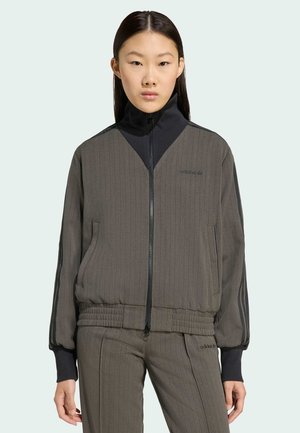 Giacca grigia a costine con zip e collo alto, con dettagli neri, tasche laterali e fascia aderente in vita. Abbinata a pantaloni coordinati.