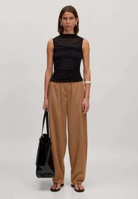 Zwarte mouwloze top met geplooide textuur, hoge halslijn; tan kleurige wijde broek; zwarte handtas; minimalistische zwarte slippers; gladde oppervlakken.