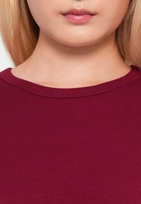 Burgundy ribbed toppi pyöreällä pääntiellä; kangas vaikuttaa pehmeältä ja sileältä. Lähikuva, joka keskittyy kauluksen ja kankaan yksityiskohtiin.