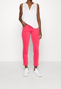 Bílá bezrukávová blůza s skládacím výstřihem, kombinovaná s jasně růžovými skinny džíny a bílými teniskami. Jednoduchý design, hladká textura.