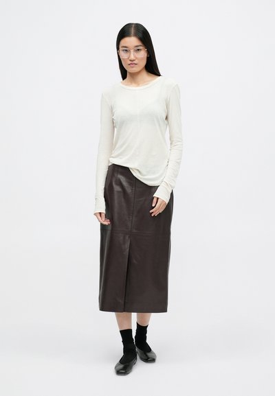 Blusa de manga larga color marfil combinada con una falda de cuero marrón oscuro con una abertura frontal, llevada con calcetines negros y zapatos negros.