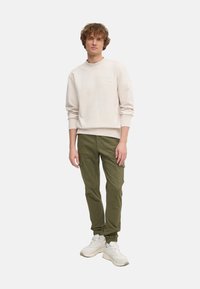 Felpa beige chiaro con collo rotondo e logo sottile, abbinata a pantaloni cargo verde oliva con tasche laterali e fasce a fondo gamba. Sneakers bianche.