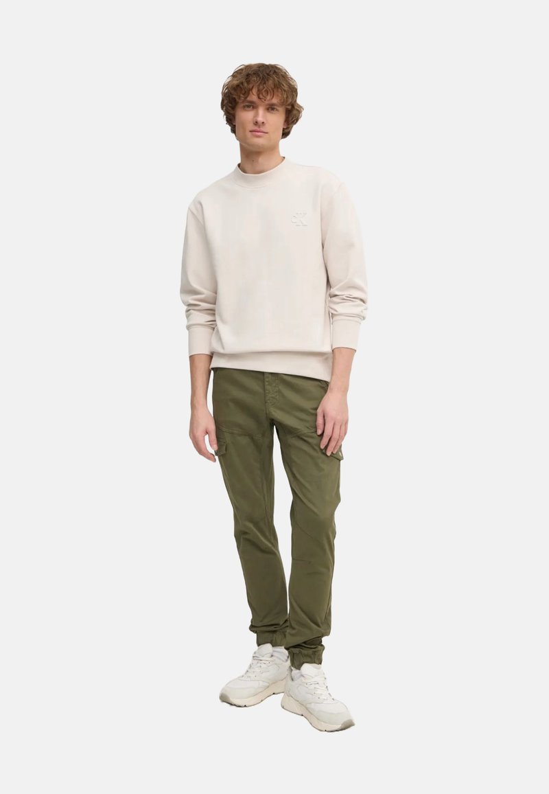 Felpa beige chiaro con collo rotondo e logo sottile, abbinata a pantaloni cargo verde oliva con tasche laterali e fasce a fondo gamba. Sneakers bianche.