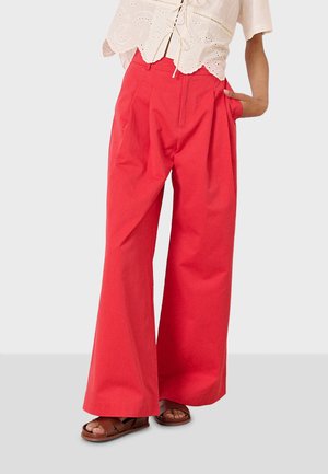 Pantalons larges rouges en tissu léger, dotés de plis à l'avant et de poches latérales. Associés à un chemisier de couleur claire et des sandales marron.
