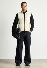 Ανδρικό γιλέκο σε ανοιχτό μπεζ χρώμα πάνω από μαύρο πουκάμισο με μακριά μανίκια, σε συνδυασμό με σκούρες παντελόνες σε φαρδιά γραμμή και μαύρα παπούτσια, κρατώντας μια μαύρη τσάντα.