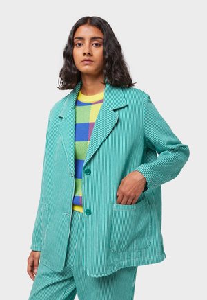 Giacca verde a righe con colletto a punte, bottoni frontali e tasche laterali, abbinata a un maglione a fantasia multicolore.