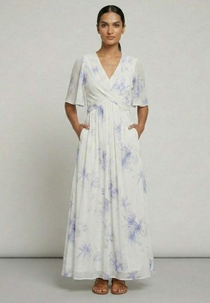 Femme debout, mains dans les poches, portant une robe longue blanche avec un imprimé floral violet clair, col en V et manches volantées, associée à des sandales.