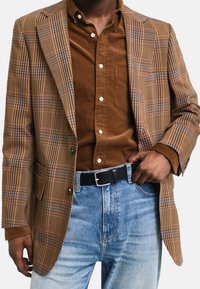 Brauner kariertes Sakko mit blauen und orangefarbenen Akzenten, kombiniert mit einem braunen Cordhemd und hellblauen Jeans, ergänzt durch einen schwarzen Gürtel.