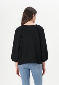 Black textured blouse with balloon sleeves, scalloped hem, and floral embroidery details at the cuffs, worn with light blue jeans.