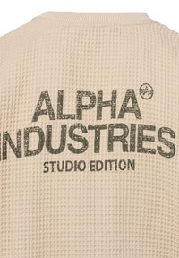 Beige textured sweatshirt met een wafelbreipatroon, voorzien van donkere groene "ALPHA INDUSTRIES" tekst en "STUDIO EDITION" eronder.