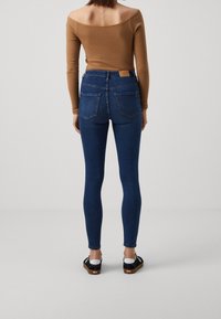 Úzké modré džíny s vysokým pasem, vybavené zadními kapsami a koženou visačkou, doplněné černými nazouvacími botami.