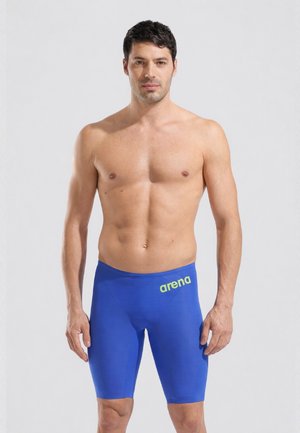 Man zonder shirt met kort donker haar, gekleed in blauwe Arena zwemjammers, staand tegen een effen lichtgrijze achtergrond.