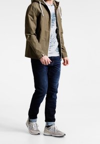 Oliven grøn letvægtsjakke med hætte, hvide snore; grå t-shirt med blå bladmønster; mørkeblå jeans; lys grå sneakers.