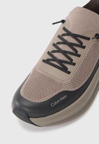 Calvin Klein sneakers med en beige mesh-overdel og såle i sort gummi; har sorte snørebånd og logo detaljer på siden. Rund tå-design.