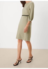 Olive green dress with three-quarter sleeves, belted at the waist with a black band. Smooth texture, knee-length hem, paired with black heels.