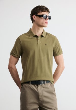 Νεαρός άνδρας με πολο σε ελαιώδες πράσινο, μπεζ παντελόνι, μαύρη ζώνη και μαύρα γυαλιά ηλίου, στέκεται με τα χέρια στις τσέπες μπροστά από λευκό φόντο.