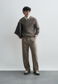 Maglione di lana marrone con scollatura a V, abbinato a pantaloni beige a pieghe. Borsa a tracolla nera e scarpe bicolore.