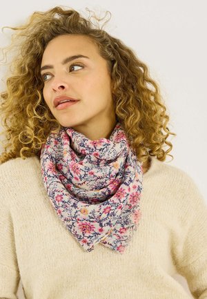 Mujer con cabello rubio rizado que lleva un suéter crema y una bufanda con estampado floral, mirando ligeramente hacia un lado sobre un fondo liso.