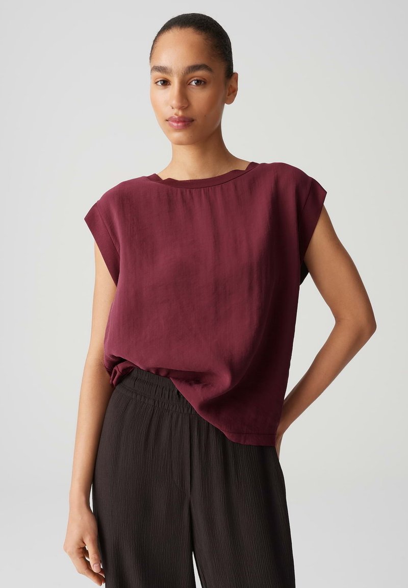 Top borgogna loose-fit con scollo rotondo e maniche corte, realizzato in un tessuto morbido, abbinato a pantaloni neri plissettati con texture.
