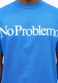 Άτομο που φοράει φωτεινό μπλε μπλουζάκι με μεγάλο άσπρο κείμενο "No Problemo" στο ύψος του στήθους.