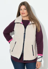 Fleece-Weste in Creme mit marineblauen Akzenten und Reißverschluss. Verfügt über zwei Seitentaschen und einen Kragen. Getragen über einem weinroten Oberteil.