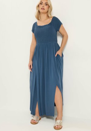 Vrouw draagt een blauwe maxi-jurk met korte mouwen, een gerimpeld lijfje, zijslitsen en zakken, gecombineerd met gevlochten slip-on sandalen.