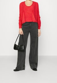 Czerwony dzianinowy kardigan na czerwonym topie, zestawiony z ciemnoszarymi, szerokimi spodniami z wysokim stanem. W prawej ręce trzymana jest mała czarna torebka.