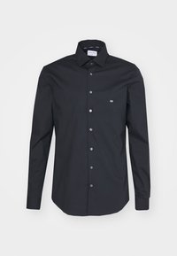 Camicia nera a maniche lunghe con bottoni, colletto a punte, piccolo logo bianco "CK" sul petto e etichetta Calvin Klein visibile all'interno del colletto.