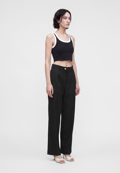 Νεαρή γυναίκα στέκεται φορώντας μαύρο crop top με λευκή λεπτομέρεια, μαύρο ψηλόμεσο παντελόνι με φαρδιά μπατζάκια και λευκές ανοιχτές κάνει.