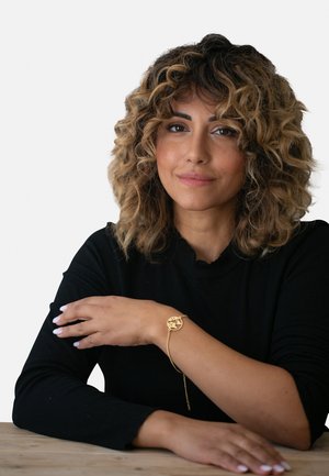 Bracelet en or avec un design de pendentif circulaire, présentant un motif découpé. Porté au poignet d'une personne vêtue d'un haut noir, montrant une peau lisse.